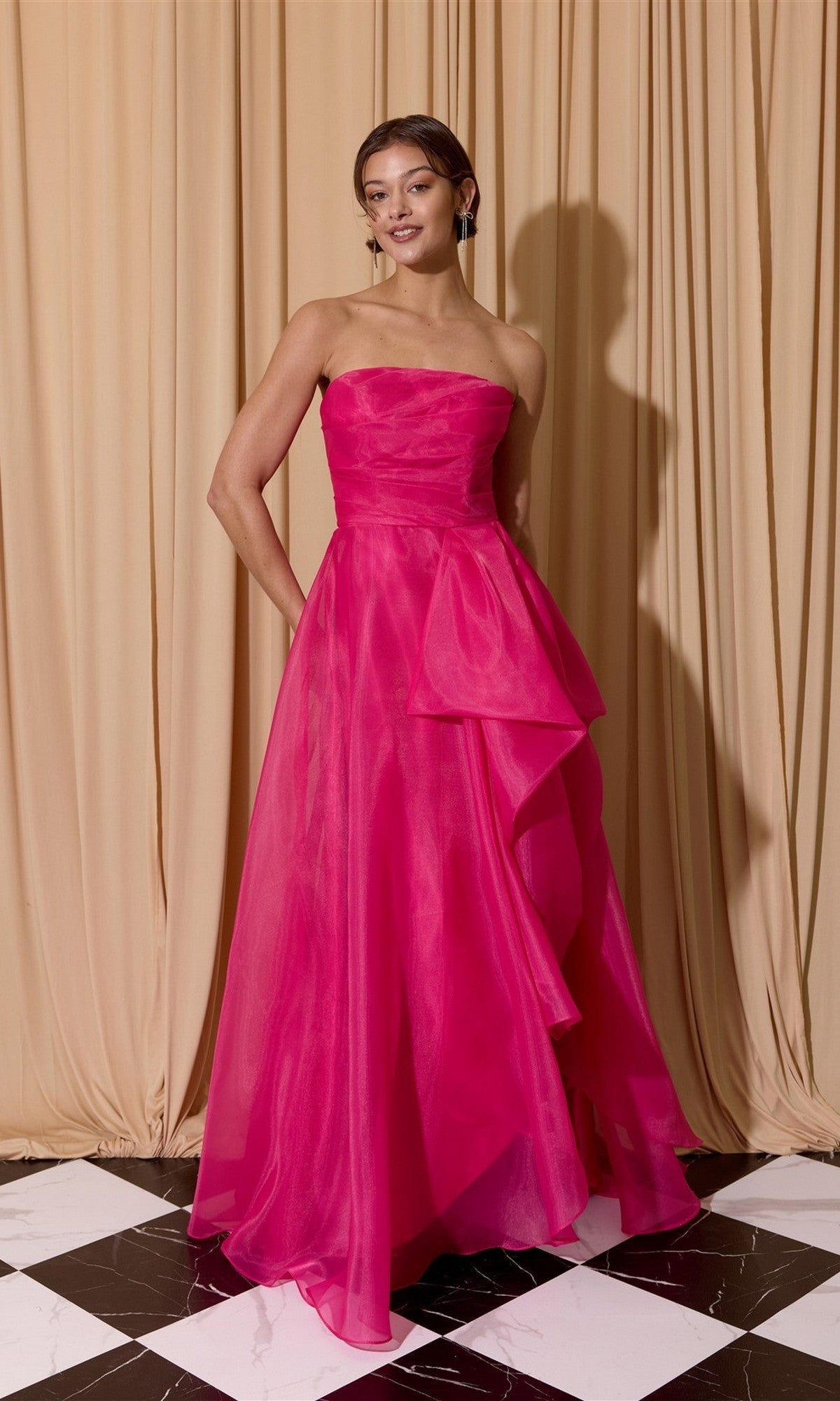 A woman stands on a black and white checkered floor wearing the Eureka EK206 long prom dress—a bright pink strapless organza gown with draped detail—in front of beige curtains.